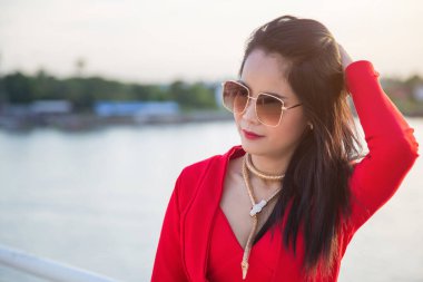 Beyaz bir çitin yanındaki kırmızı ceketli Asyalı kadını kapat. Sol eliyle köprünün arkasında nehir altyapısı olan saçına dokunuyor.