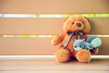 Kahverengi oyuncak ayı tahta bir bankta oturuyor, bacağında sincap bebeği oturuyor, sol tarafı boş.