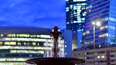 Geceleri şehir arkaplanlı çeşme manzarası. Şehir çeşmesine karşı cam modern binalar geceleri arka planda kalıyor. Modern bir cephenin mimari ızgarasından çıkan bulanık pencereler. 