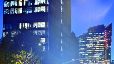 Şehir merkezindeki modern ofis binası gece aydınlandı. Modern bir ofis binasının yanındaki yeşil ağaçların önünde cam bir cephe var. Modern bir cephenin mimari ızgarasına karşı yanan pencereler.