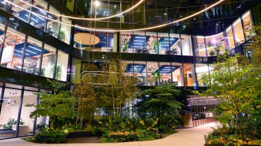 Şehir merkezindeki modern ofis binası gece aydınlandı. Modern bir ofis binasının yanındaki yeşil ağaçların önünde cam bir cephe var. Modern bir cephenin mimari ızgarasına karşı yanan pencereler.