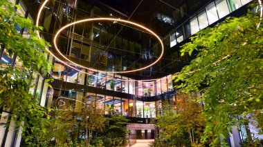 Şehir merkezindeki modern ofis binası gece aydınlandı. Modern bir ofis binasının yanındaki yeşil ağaçların önünde cam bir cephe var. Modern bir cephenin mimari ızgarasına karşı yanan pencereler.