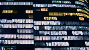 Şehir merkezindeki modern ofis binası gece aydınlandı. Aydınlatılmış pencereler modern bir cephenin mimari şebekesine karşı. Gece versiyonunda beton ve cam cephe.