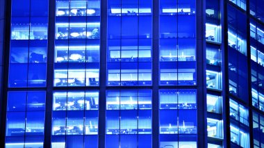 Modern ofis binasında geceleri büyük parlak pencereler, pencereler ışık saçıyor. Geceleri modern bir şirket binasının cam cephesinin bir parçası. Şehirde modern cam ofis.