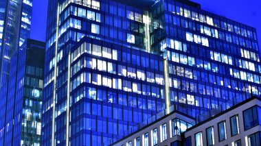 Modern ofis binasında geceleri büyük parlak pencereler, pencereler ışık saçıyor. Geceleri modern bir şirket binasının cam cephesinin bir parçası. Şehirde modern cam ofis.