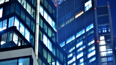 Modern ofis binasında geceleri büyük parlak pencereler, pencereler ışık saçıyor. Geceleri modern bir şirket binasının cam cephesinin bir parçası. Şehirde modern cam ofis.