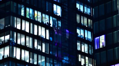 Modern ofis binasında geceleri büyük parlak pencereler, pencereler ışık saçıyor. Geceleri modern bir şirket binasının cam cephesinin bir parçası. Şehirde modern cam ofis.