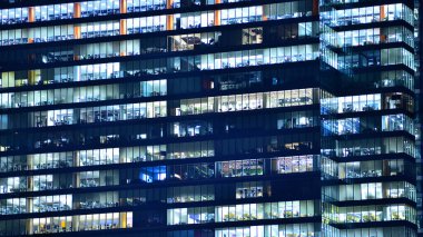 Modern ofis binasında geceleri büyük parlak pencereler, pencereler ışık saçıyor. Geceleri modern bir şirket binasının cam cephesinin bir parçası. Şehirde modern cam ofis.