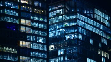 Modern ofis binasında geceleri büyük parlak pencereler, pencereler ışık saçıyor. Geceleri modern bir şirket binasının cam cephesinin bir parçası. Şehirde modern cam ofis.