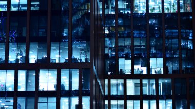 Modern ofis binasında geceleri büyük parlak pencereler, pencereler ışık saçıyor. Geceleri modern bir şirket binasının cam cephesinin bir parçası. Şehirde modern cam ofis.