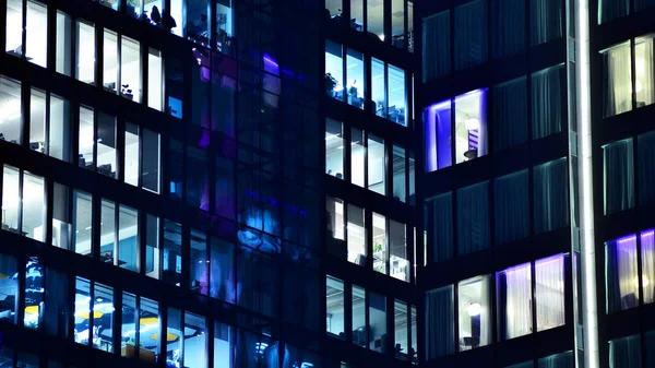 Modern ofis binasında geceleri büyük parlak pencereler, pencereler ışık saçıyor. Geceleri modern bir şirket binasının cam cephesinin bir parçası. Şehirde modern cam ofis.