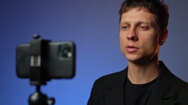 Takım elbiseli genç adam RGB renkli arka planlı akıllı telefon tripodunda motivasyon videosu kaydediyor..