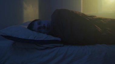 Genç adam geceleri gözleri açık bir şekilde uzanıyor. Ön plandaki bulanık saat ve sıcak pencere ışığı uykusuzluk atmosferi yaratır.