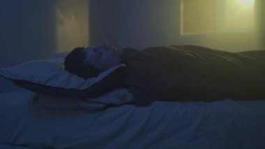 Genç adam geceleri gözleri açık bir şekilde uzanıyor. Ön plandaki bulanık saat ve sıcak pencere ışığı uykusuzluk atmosferi yaratır.