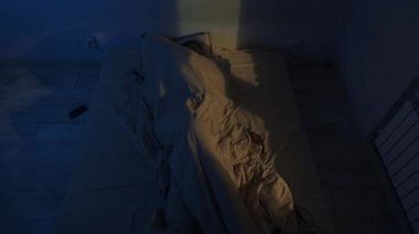 Genç adam geceleri gözleri açık bir şekilde uzanıyor. Ön plandaki bulanık saat ve sıcak pencere ışığı uykusuzluk atmosferi yaratır.