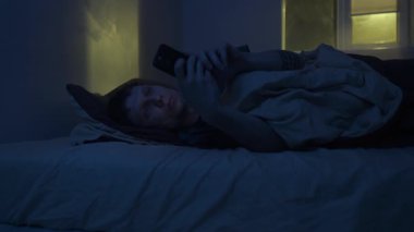 Yorgun bir adam karanlık yatak odasında uyanık, kaydırmalı telefonda yatıyor. Mavi ekran ışığı uykuyu engeller, uykusuzluk, endişe ve dijital bağımlılık gösterir.