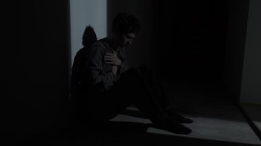 Karanlık bir odada yerde oturan bir adam panik atak sırasında göğsünü sıkıca tutuyor. Gerçek endişe, korku, zihinsel sağlık krizi, duygusal acı.