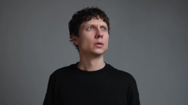 Siyahlı genç adam başını çeviriyor ve evde şüpheli bir ses dinliyor. Endişe, paranoya, ev güvenliği ve tarafsız gri arka planda davetsiz misafir korkusu..