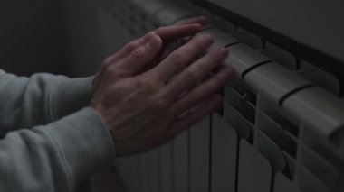 Yüksek enerji maliyetlerinden dolayı soğuk, soğuk dairede radyatörün yanında ellerini ısıtan adam ısıtma faturaları ve enerji sıkıntısı ile mücadele ediyor.