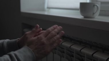 İnsani kriz konsepti, soğuk dairede radyatörün yanında elini ısıtan adam, enerji kıtlığı ve ısıtma sistemi hatası, küresel istikrarsız yaşam tarzı