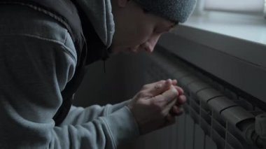 Yüksek enerji maliyetlerinden dolayı soğuk, soğuk dairede radyatörün yanında ellerini ısıtan adam ısıtma faturaları ve enerji sıkıntısı ile mücadele ediyor.