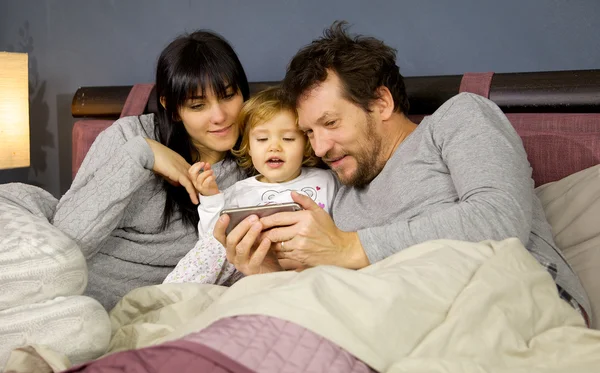 Aranyos boldog család az ágyban néz video a mobiltelefon közepes kislányom lövés — Stock Fotó