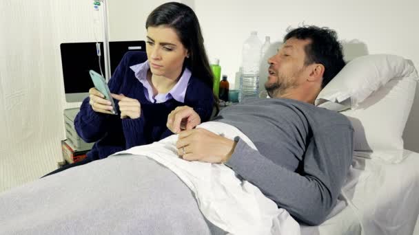 Чоловік у лікарняному ліжку сміється, дивлячись фотографії на телефон з дівчиною — стокове відео