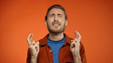 bearded young man with closed eyes and crossed fingers on orange  clipart