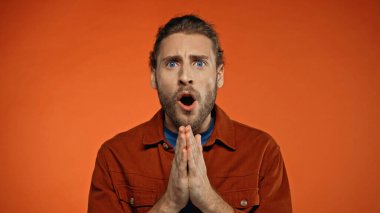 shocked young man with praying hands looking at camera on orange  clipart