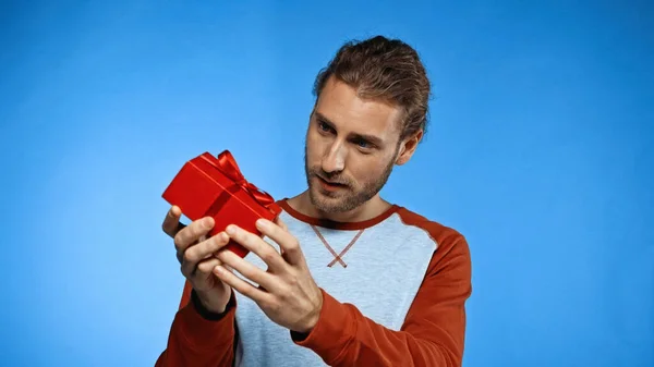 Nieuwsgierige Jongeman Kijkt Naar Verpakt Rood Cadeau Blauw — Stockfoto