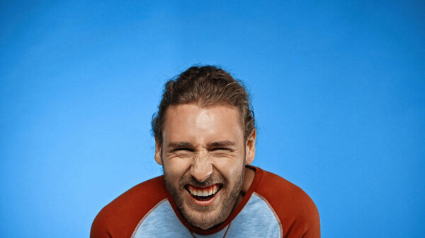 bearded young man looking at camera and laughing on blue