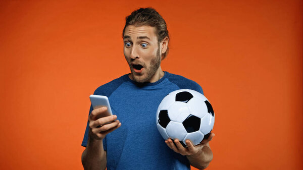 shocked football fan in blue t-shirt using smartphone and holding soccer ball on orange 