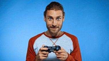 KYIV, UKRAINE - NOVEMBER 24, 2020: excited man holding joystick while playing video game on blue  clipart