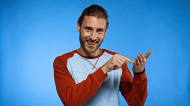 excited young man pointing with finger at hand while looking at camera on blue clipart