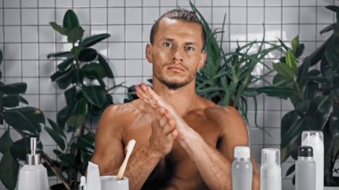 Tişörtsüz bir adam banyoda arka planda yeşil bitkilerin yanında krem sürüyor. 
