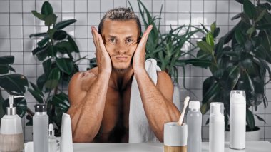 Üstsüz bir adam banyoda yüzüne dokunuyor. Arka planda bitkiler var.