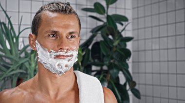 Yüzünde tıraş köpüğü olan gömleksiz bir adam bulanık arka plandaki bitkilerin yanından uzağa bakıyor.