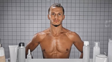 Banyoda şişelerin yanında duran kaslı ve sakallı adam. 