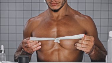 partial view of shirtless man holding wax strip on chest in bathroom  clipart