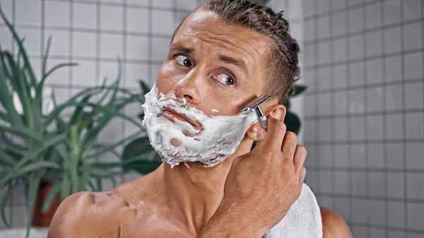 man with shaving foam on face holding razor with plants on blurred background