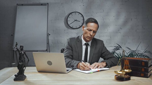 Insurance agent in suit writing on notebook near laptop, books and scales on table 