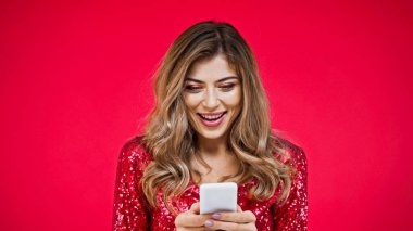 amazed woman with wavy hair texting on smartphone isolated on red clipart