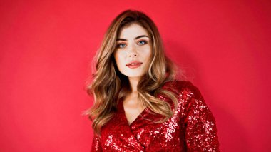 Kıvırcık saçlı ve mavi gözlü bir manken kırmızıda kameraya bakıyor.