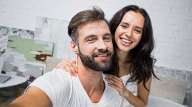 Mutlu sakallı adam ve esmer kadın yatak odasında kameraya gülümsüyor.