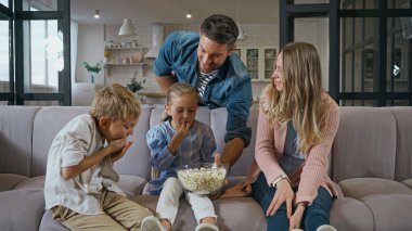 Çocuklar koltukta gülümseyen ebeveynlerin yanında patlamış mısır yiyorlar. 