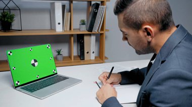 Yeşil ekranlı dizüstü bilgisayar. İş adamının yanında. Defterde bulanık önplan yazıyor. 