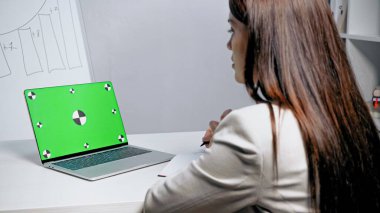 Ofisteki bulanık önplanda iş kadınının yanında yeşil ekranlı bir laptop. 