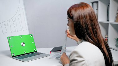 İş kadınının yanında yeşil ekranlı dizüstü bilgisayar ofiste bulanık önplanda belge gösteriyor 