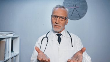 Hastanenin bulanık ön planında elleri kamerada gülümseyen doktor. 