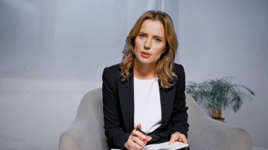 Yetişkin bir iş kadını elinde kalem ve defterle kamerayı işaret ediyor. 